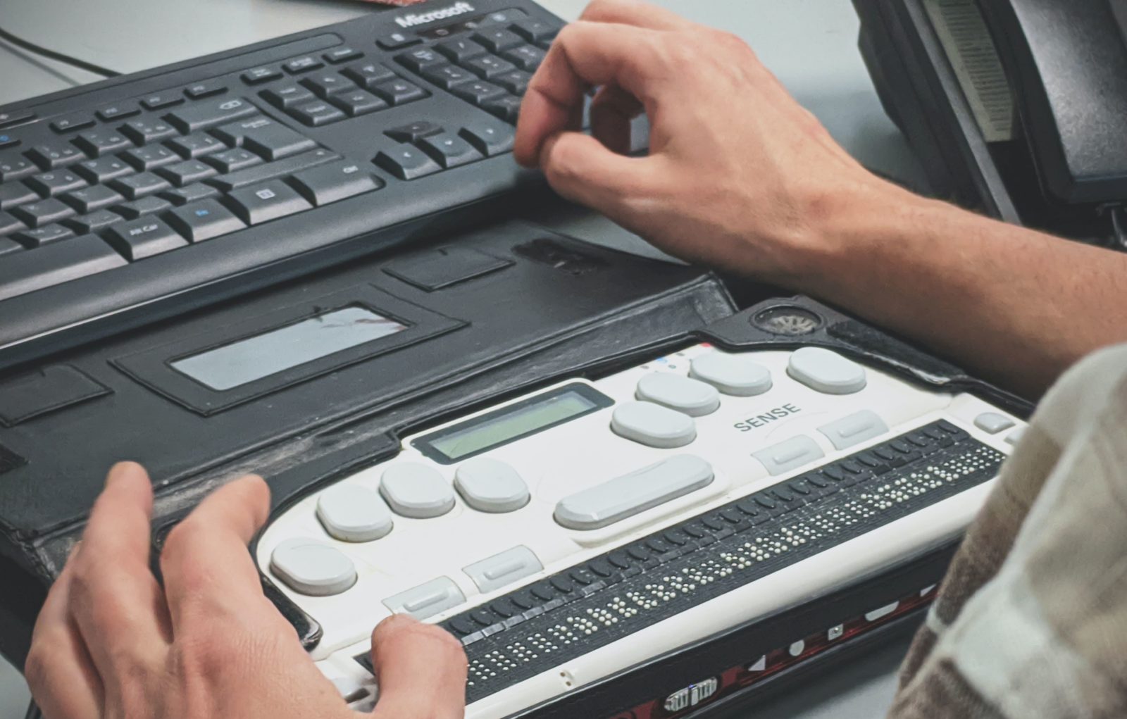 A man uses his right hand to type into a keyboard while his left hand hovers over a screen reader device.