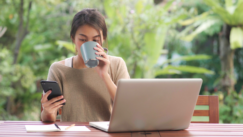 Woman in a brown sweater drinking coffee and looking at her phone in front of a laptop computer