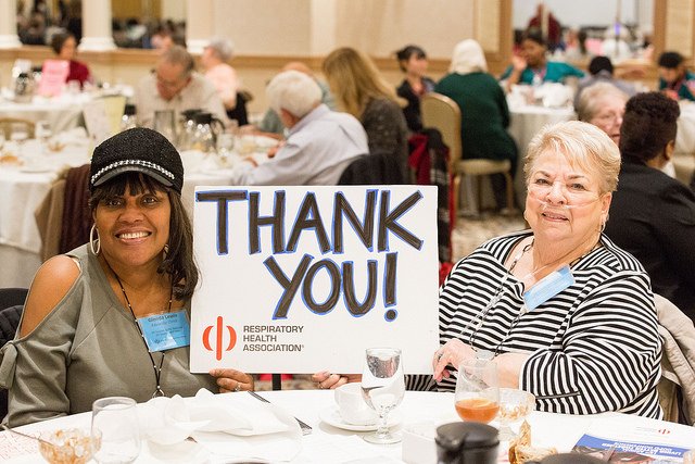 Two women are sitting at a table at a nonprofit luncheon. They hold between them a sign that says "Thank You!" in large, handwritten letters. In the bottom left corner of the sign is the Respiratory Health Association logo.