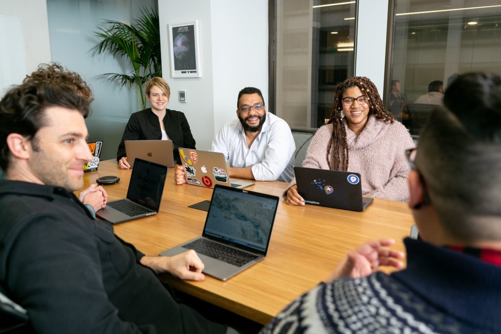 A group of young adults sit around a conference table. Each person has an open laptop in front of them.