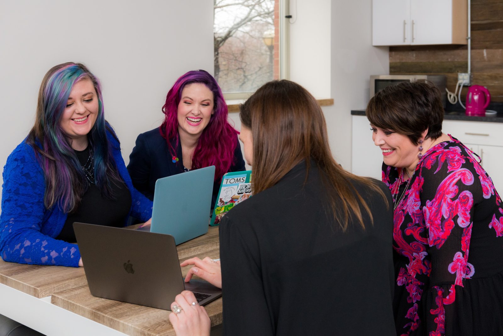 4 women sit around an office table smiling and working on their laptops