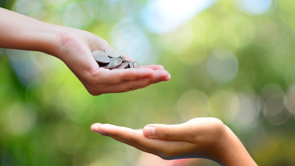 A comprehensive donor management software comparison can help you find the right program for your cause. In this image, two hands appear in front of a blurred background in shades of green and white. One cupped hand is full of coins; below it, another outstretched hand is ready to receive that money.