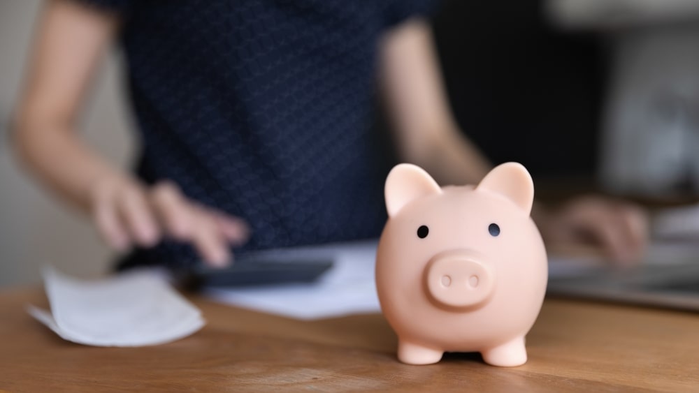 Knowing how to calculate the donor lifetime value can help your nonprofit grow and thrive. This image depicts a matte pink piggy bank that seems to be looking into the camera. In the background, someone wearing a dark-blue shirt is working with a calculator and laptop.