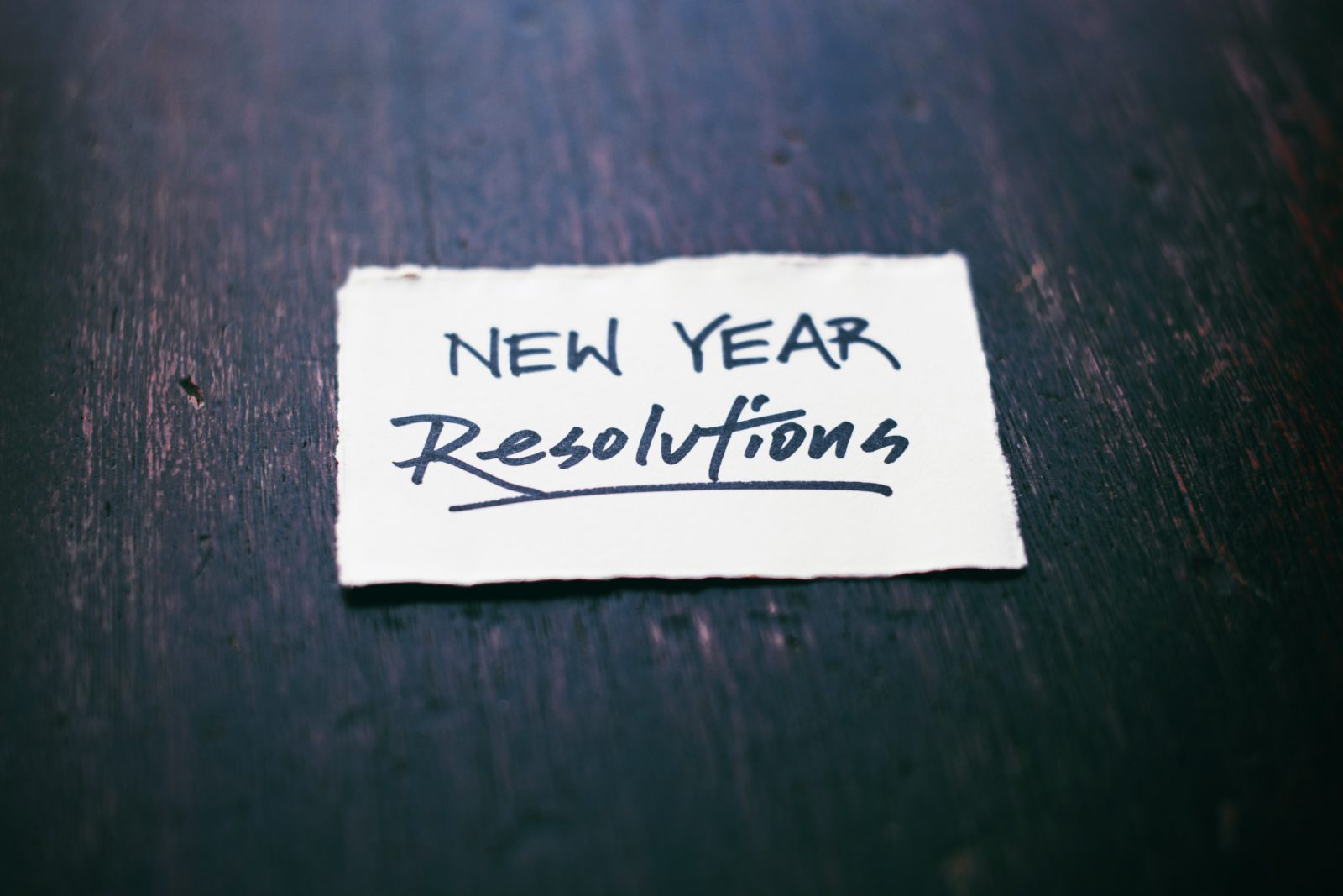 A square slip of paper rests on a dark wooden table. The words "New Year Resolutions" are written on the paper in heavy black ink.