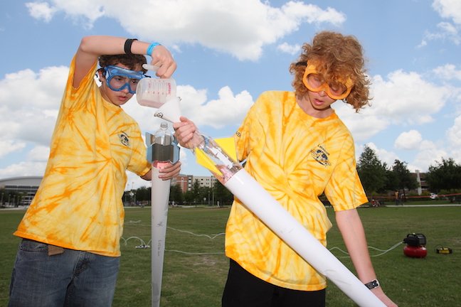 Two Science Olympiad participants in bright yellow shirts and protective gear are working with bottle rockets they built for the program. They're standing in a sunny field preparing to launch their craft.
