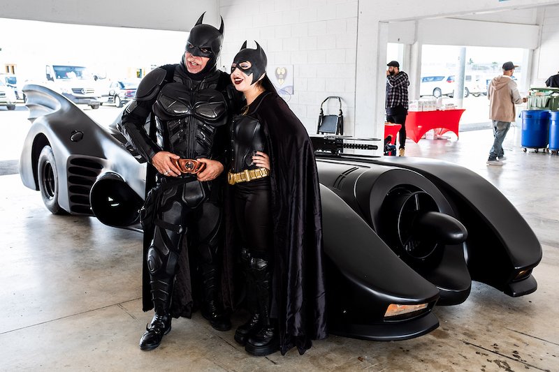 Two people dressed as Batman and Batgirl standing in front of the Batmobile