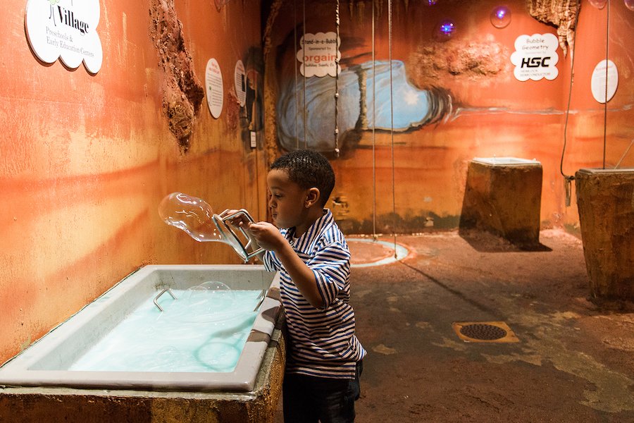 Little boy blowing a giant bubble over a tub of soap water in a red-colored room.