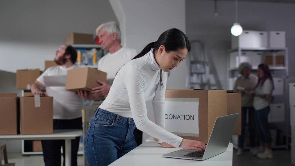 Donation management software can help you track donations of all types while building constituent relationships. In this image, a woman works on laptops in a warehouse where donations are collected.