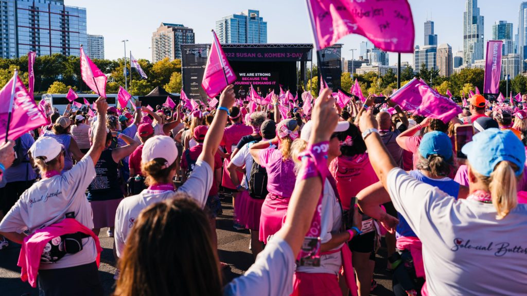 A large group of people wearing pink and waving pink flags cheer at an outdoor fundraiser.