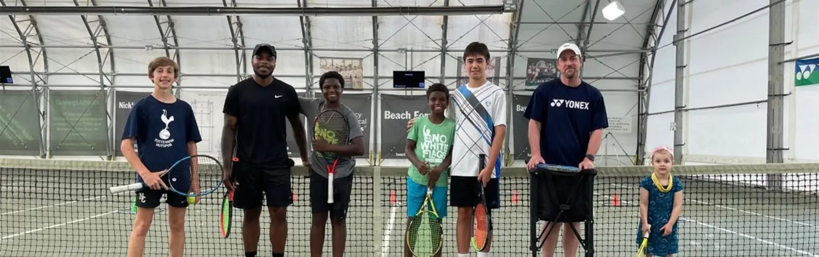 Group of 7 people of mixed ethnicities and ages holding tennis rackets standing in front of a tennis ball net.