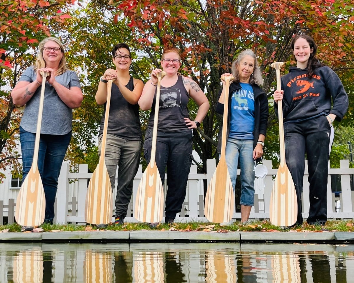 Five students displaying their wooden paddles at the edge of a pond.