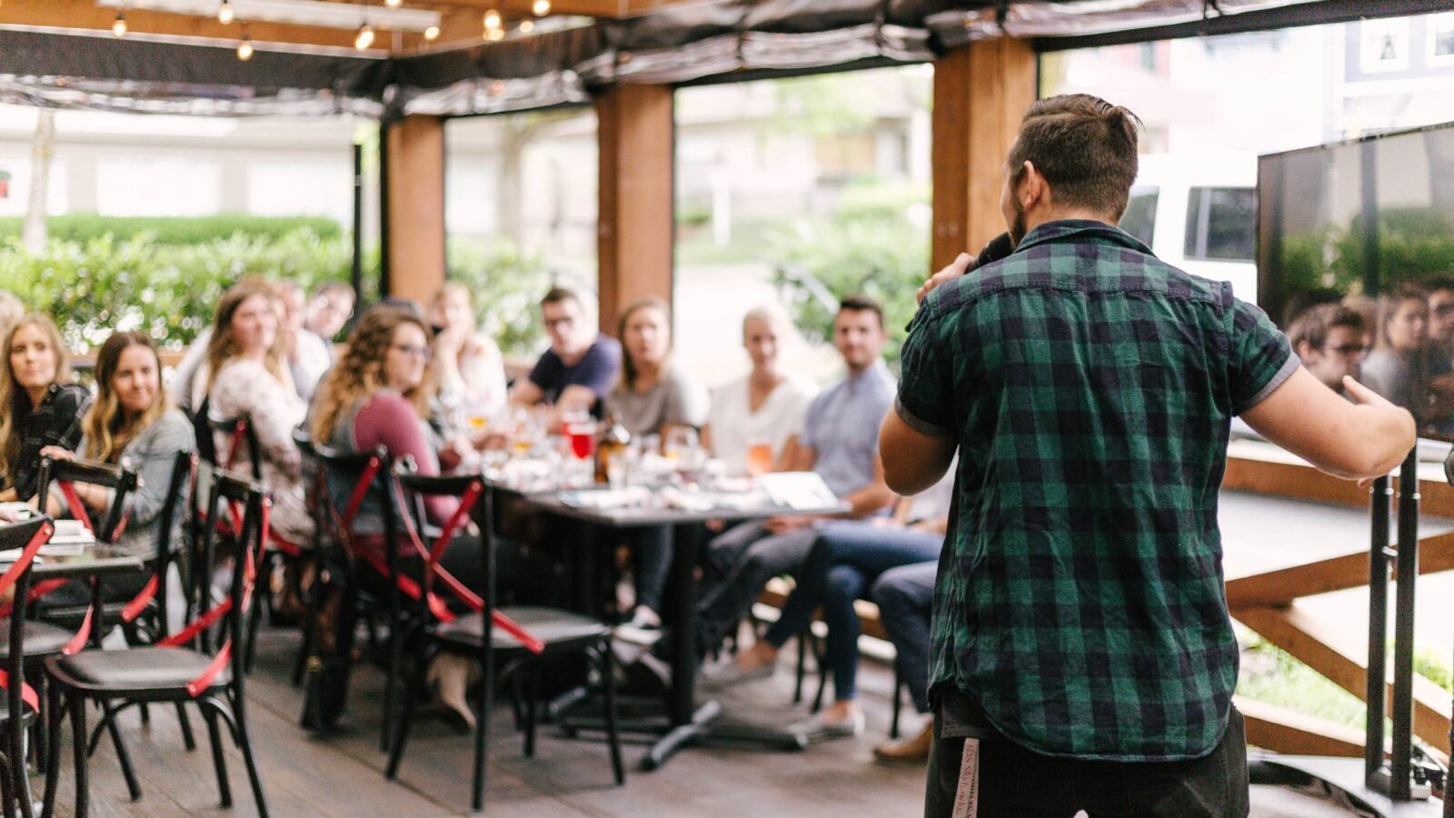 in this photo, a man is speaking into a microphone in front of people seated at long tables during a fun nonprofit event