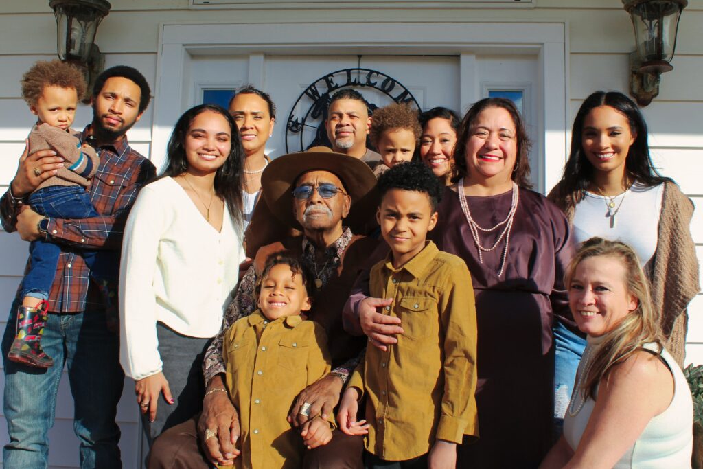 This photo shows several generations of a family. There are 12 people of all ages in the photo, and all of them are gathered together in front of their home.