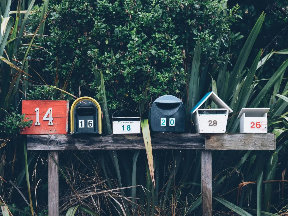 6 mailboxes of differnt sizes and colors sitting on a wooden board surrounded by lush greenery.